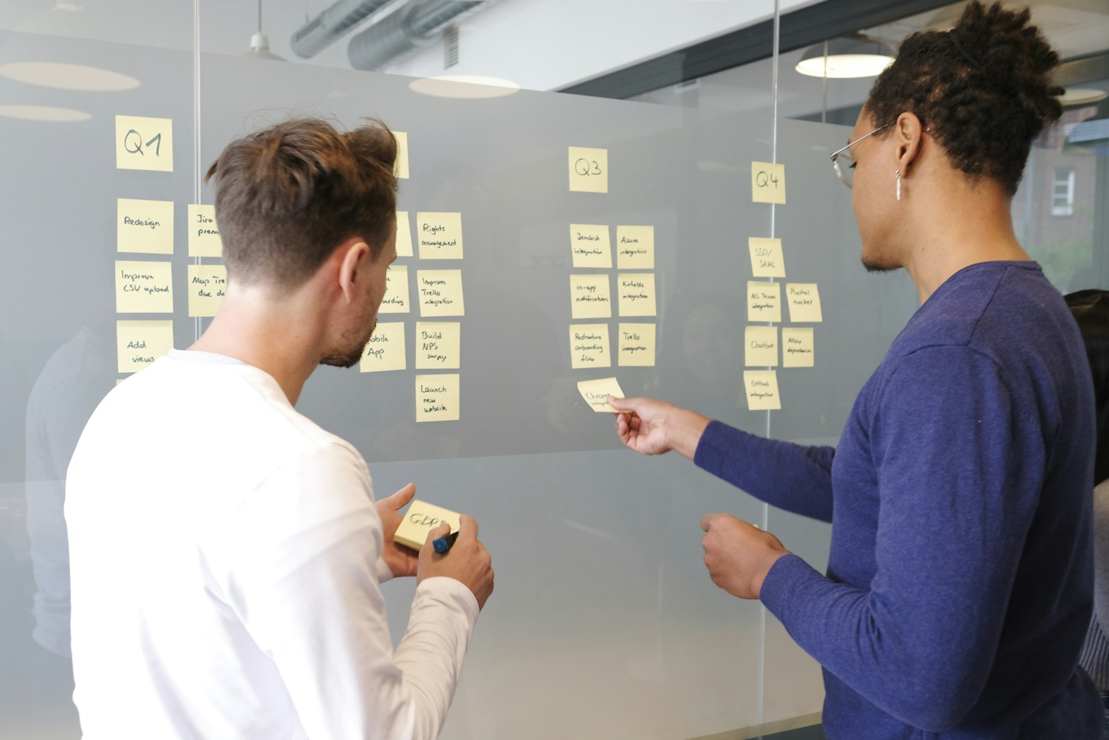 Two people collaborating on a project plan using yellow sticky notes on a glass wall