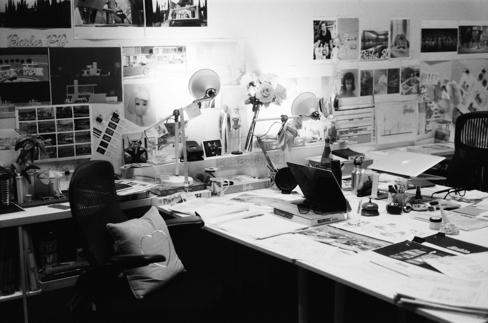 Messy workspace with desk, papers, lamps, and photos on wall