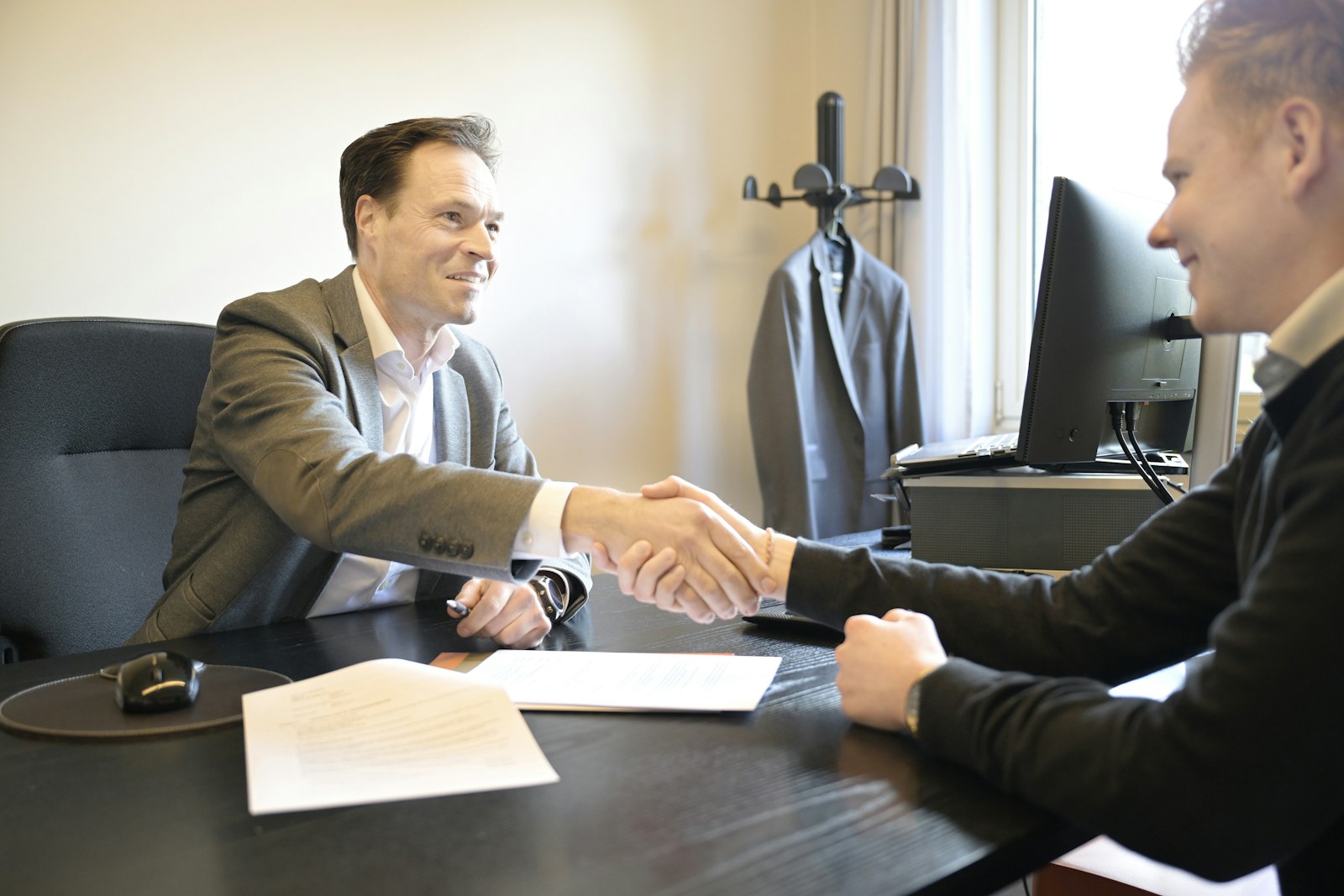 Two smiling men shake hands across an office desk
