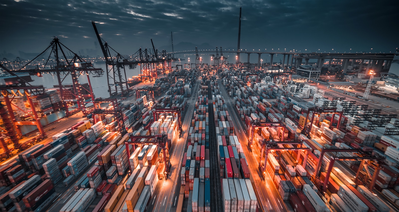 A busy shipping port with stacked containers and cranes under a dark cloudy sky