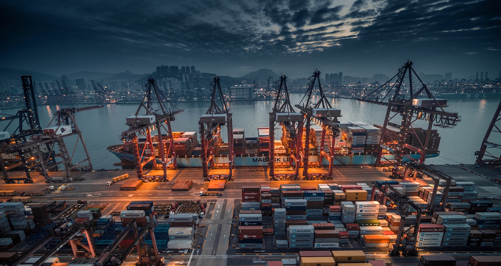 A container ship being loaded by cranes at a busy industrial port at dusk