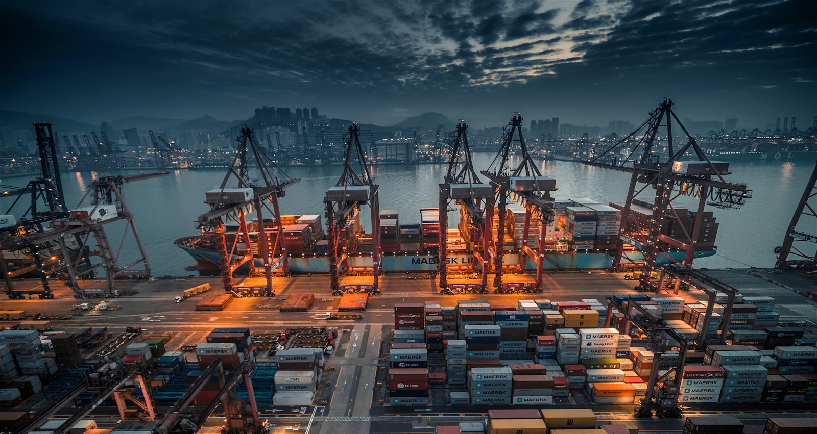 A container ship being loaded by cranes at a busy industrial port at dusk