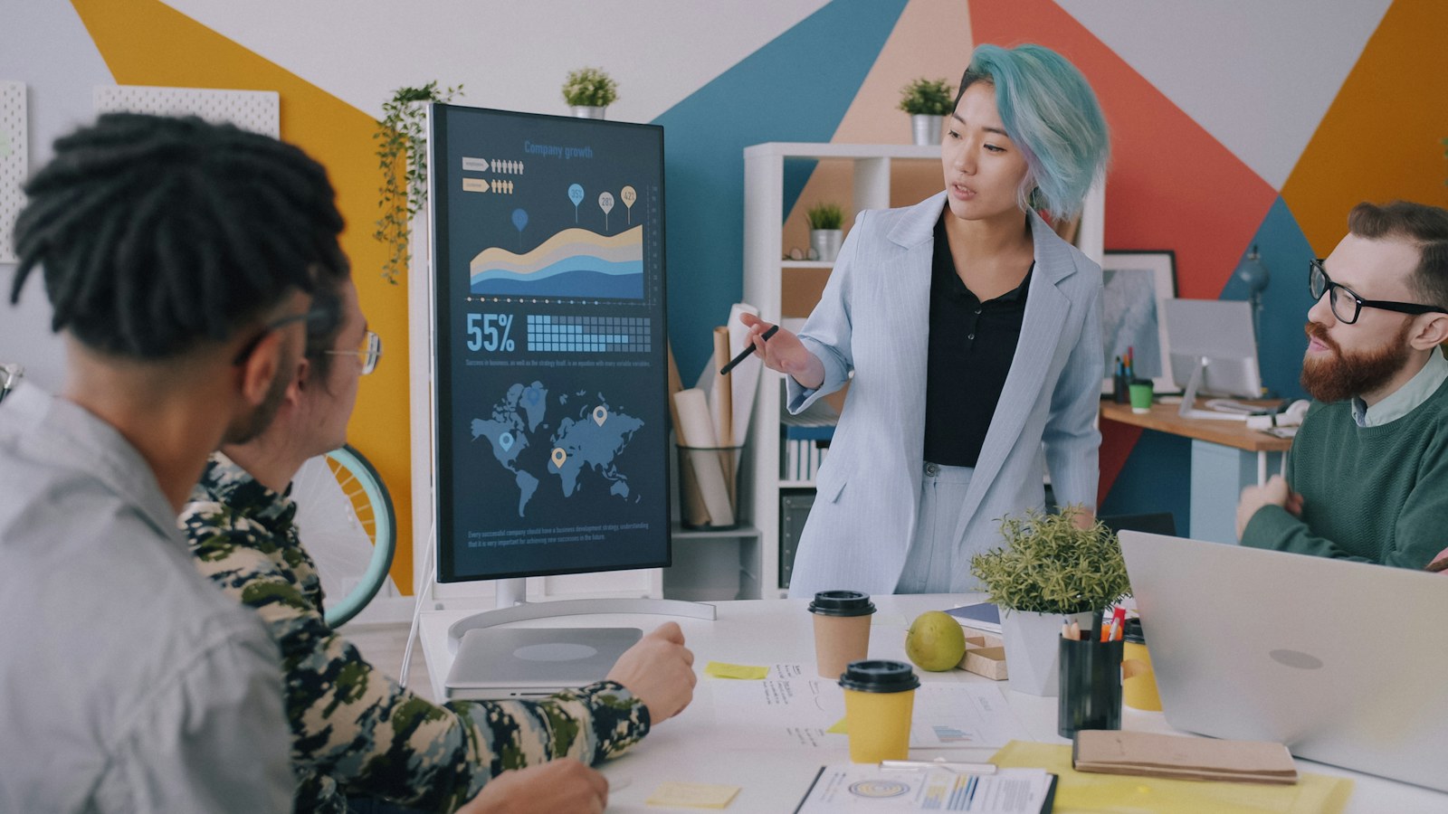 A woman with blue hair presenting data on a monitor to a team
