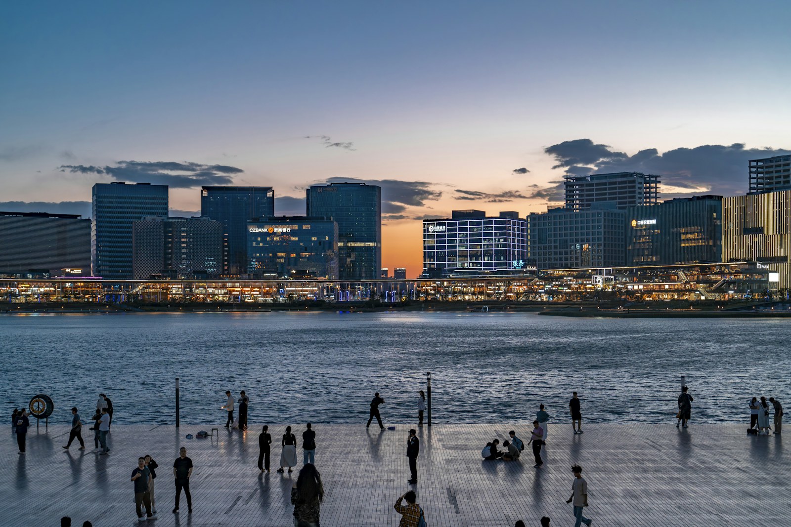 People on a waterfront promenade at dusk with a modern city skyline behind